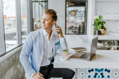 Junger Mann in Blazer sitzt an Tisch mit Laptop und Getränk, schaut nachdenklich aus dem Fenster.